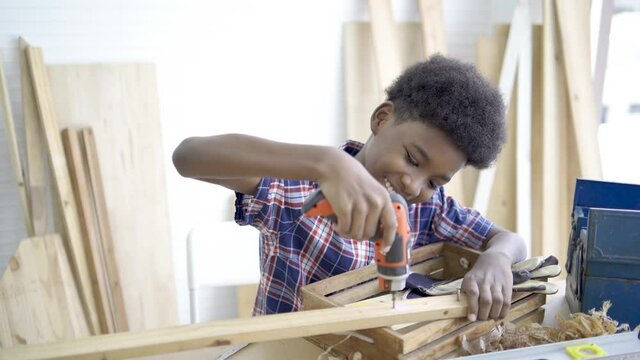 A young african boy wearing shirt and using screwdriver and try to screw the wood piece in the classroom. Smiling kid carpenter happy working with wood and sandpaper, preschool and learning concept.