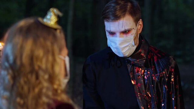 Portrait of young man in vampire costume with face paint in coronavirus face mask celebrating Halloween night with little girl in dark forest. Scary Caucasian father having fun with daughter on Covid