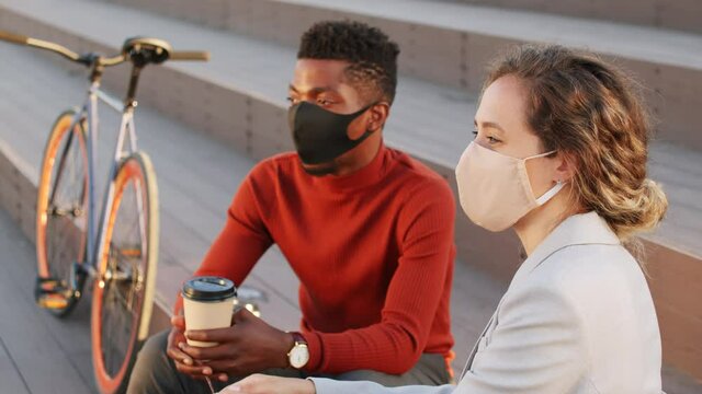 Medium slowmo shot of young successful interracial business partners wearing face masks talking about work sitting on stairs outdoors on sunny day