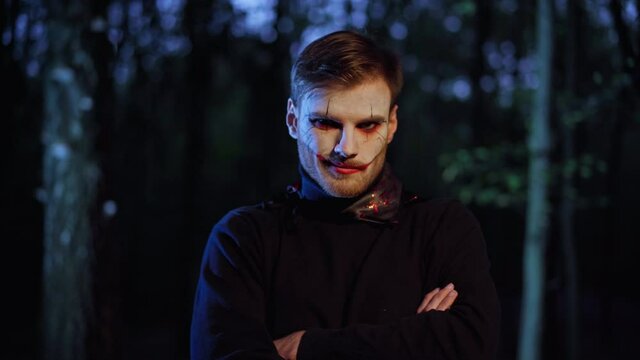 Spooky scary man with vampire face paint standing in night forest looking at camera on Halloween. Portrait of young Caucasian guy posing in woods outdoors on holiday. Leisure and celebration