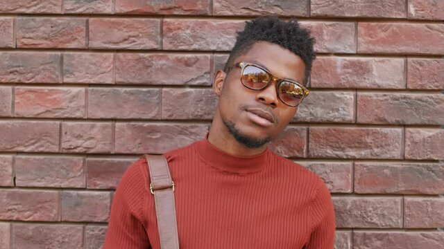 Medium close-up portrait with slowmo of handsome young African-American man wearing red turtleneck and trendy sunglasses posing for camera standing against brick wall outdoors