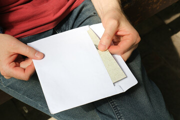The man opens the envelope and takes out a postcard. The young man received the letter in the envelope and will read it. White envelope in the hands of the guy.
