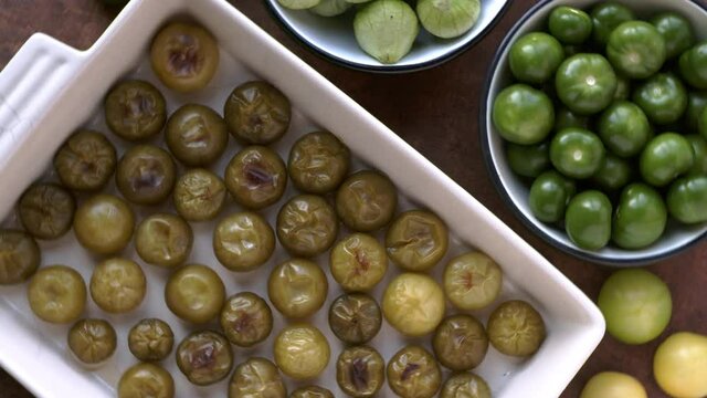 Roasted tomatillos on a baking tray and fresh green fruits in bowl. Home cooking top view rotating table.