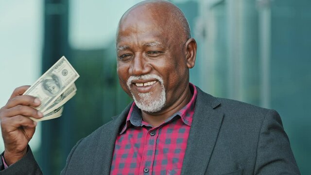 Elderly happy senior pensioner, retiree, grandfather, looking at camera, holds dollars in hands, waves fan of money to face, confident old African American man feels success, won lottery got income