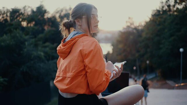 The ambitious young woman, freelancer, is working on a new project, making notes in her notebook. Dressed in a bright orange blouse, hair caught in a bun, positioned on the park strip. 4k concept