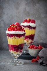 Trifles, layered dessert in glass with berry jelly, custard, sponge and whipped cream, decorated with fresh red currant and raspberry. Dark grey background, vertical image.