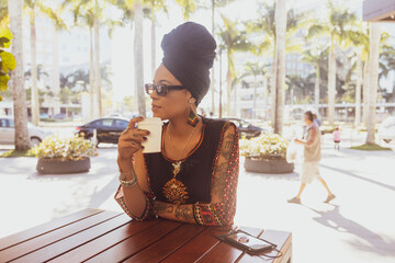 Close up of beautiful African American woman wearing sunglasses and afro turban holding a cup of coffee looking serious to the side. Mobile cell phone on the table.