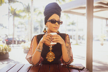 Close up of beautiful African American woman wearing sunglasses and afro turban holding a cup of coffee looking serious at the camera. Mobile cell phone on the table.