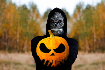 Grim reaper holding halloween pumpkin head. Man in death mask with fire flame in eyes on nature oak...