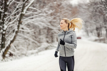 Happy sportswoman jogging on snowy path in woods at winter. Healthy lifestyle, winter fitness