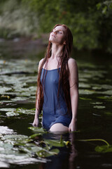Beautiful young woman with long wet hair in simple blue dress standing in water among lily pads. Sensual portrait of a girl with head up