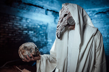 a man dressed as a plague doctor holds a human skull in his hand. costume party for Halloween....