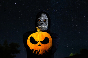 Grim reaper holding halloween pumpkin head. Man in death mask with fire flame in eyes on night sky...