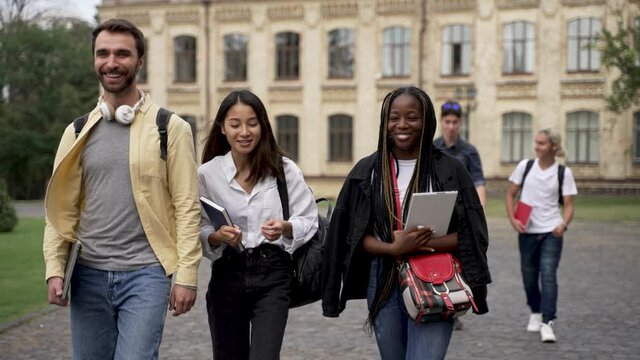 Joyful multiracial students communicating while going along park alley on background of university building. Cheerful asian and african american girls chatting with caucasian friend during walk