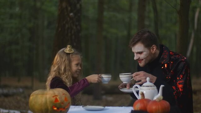 Cute charming girl clinking tea cups with man in vampire costume drinking hot drink outdoors in forest on Halloween. Caucasian kid enjoying holiday celebration with evil creature in woods