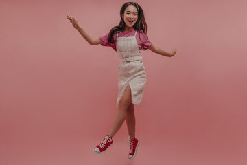 Naklejka premium Stylish young asian lady with long dark hair jumps and smiles vividly on pink background. In short white denim dress, pink T-shirt, light leather belt, and red shoes. Summer positive mood concept