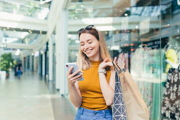 Young woman consumer in the mall browses chat and uses using a smartphone. female standing with a mobile phone in her hands in shopping center. indoor. happy shopper girl with gift bags make purchases - obrazy, fototapety, plakaty