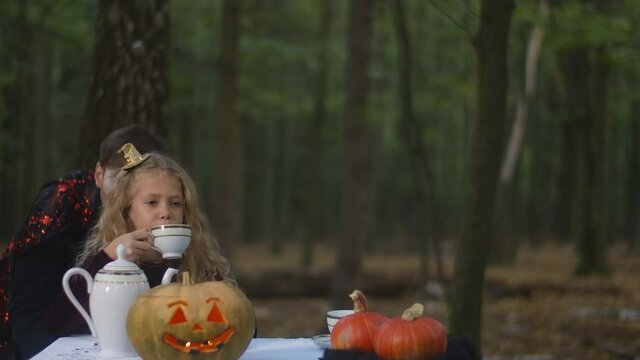 Beautiful girl sitting at table in forest drinking tea as vampire scaring kid. Pretty Caucasian child celebrating Halloween in woodland as evil entity frightening kid outdoors. Holiday concept