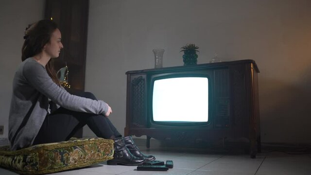 Lonely woman on isolation watching TV at home, sad female sitting on pillow on the floor and watching old retro TV with flicking screen. Boring atmospheric mood in dusk room with vintage furniture