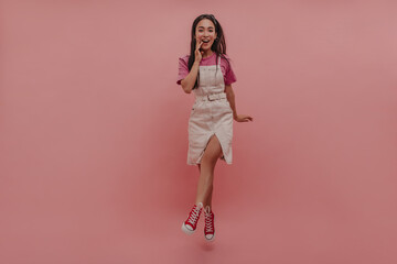 Surprised young woman from central asia jumping high and smiling happily against pink background. In short white denim dress, crimson T-shirt and red shoes, brunette in photo.