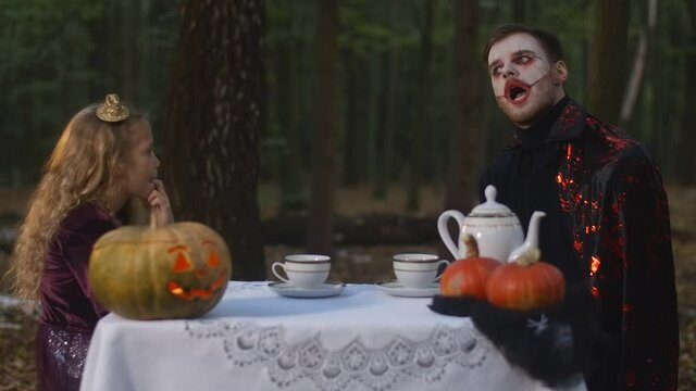 Thoughtful little girl and man in vampire costume with face paint sitting at table in forest examining each other. Caucasian kid and entity in woodland outdoors on Halloween