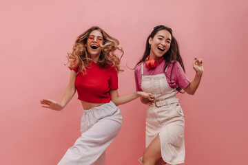 Wonderful young racial girls are dancing to beat of music on pink background in studio. Pretty women in colorful casual clothes are smiling broadly holding hands. Merry holiday concept