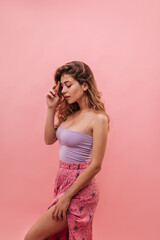 Gentle young caucasian lady from America looks down with embarrassment while straightening her hair against pink background. Medium size blonde girl posing in lovely summer skirt and purple top.