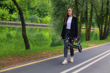 Fototapeta premium A young woman in stylish clothes walks along a bike path with a folded electric scooter