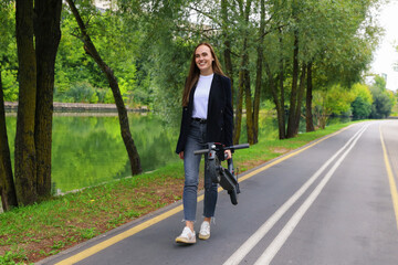 A young woman in stylish clothes walks along a bike path with a folded electric scooter