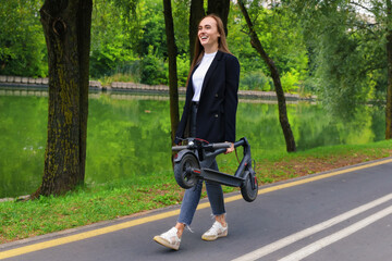 Fototapeta na wymiar A young woman in stylish clothes walks along a bike path with a folded electric scooter