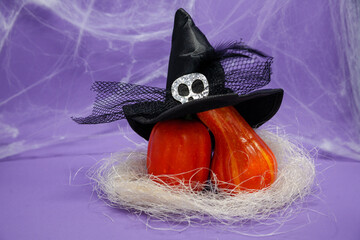 Halloween, decoration and holidays concept - Two small pumpkins of different shapes on straw leaning on each other with a witch's hat with a skull and spider web on a violet background with copy space