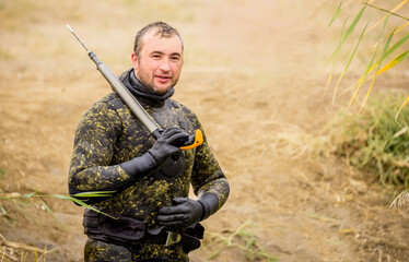 A fisherman in a diving suit stands on the bank of the river with a diving ammunition. Spearfishing, a man in a neoprene underwater suit, camouflaged clothing, a gun, diver camouflage with a harpoon.