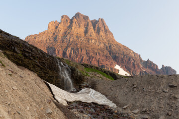 Clements Mountain catches the first of the morning's light as melting snow pours over a small waterfall towards the Hidden Lake trail in Glacier National Park Montana.	