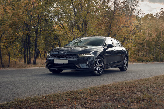 Samara, Russia - 09.12.2021: Black KIA K5 against the background of the autumn forest. The car is parked on an asphalt road near a colorful forest. Shooting a car at sunset. The front part