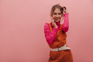 Medium shot of pretty young european blonde woman with joyful smile dancing against pink background. In orange jumpsuit with crimson long sleeve, her hands gently touch face.