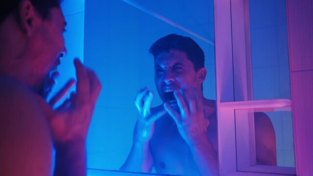Angry man with stubble on his face washes his face in the sink and screams loudly in frustration and depression while looking in the mirror in neon lighting.