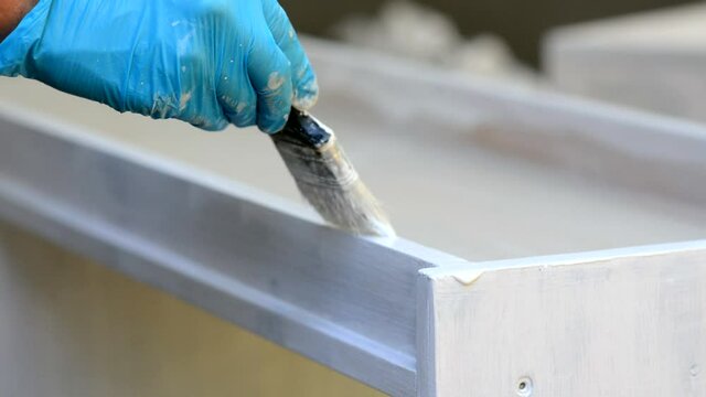 Male hands in blue protective gloves hold a paintbrush and paint a new wooden plank with white paint outdoors. The man is doing construction work. Close-up. Copy space