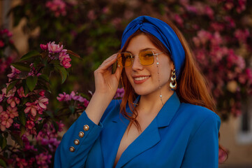 Happy smiling redhead woman wearing trendy blue turban headband, yellow sunglasses, big golden earrings, classic blazer, posing in street with blooming tree. Copy, empty space for text
