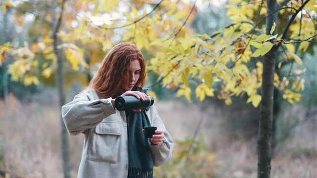beautiful red-haired woman drinks tea from thermos in the autumnal forest