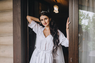 Fototapeta premium A beautiful, sexy bride in white lingerie with a long veil stands near a wooden house with a window in the morning. Wedding photography, portrait.