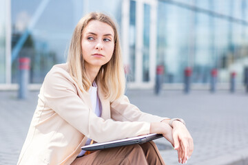 longing young woman sitting alone on street sidewalk on urban background modern building. Sad girl Student or businesswoman misses. Unemployed female lost her job, upset and lonely thoughtful, pensive