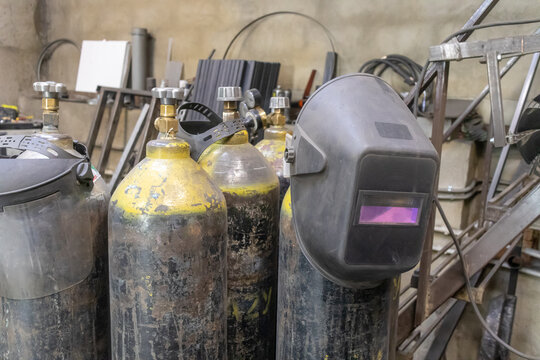 Gas cylinders for gas welding are in the workshop. The welder's mask hangs on the cylinder. Metal blanks in the background.