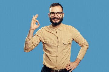 Naklejka premium Positive young male wearing eyeglasses and beige shirt showing OK gesture and looking at camera with excited face expression against blue background