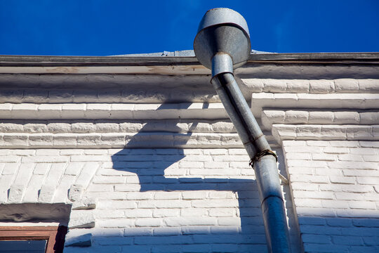 a rain gutter of a galvanized storm downspout pipe at the eaves of a building with a white facade made of white bricks on a sunny clear day.