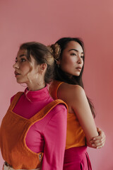 Cute young european girl stands with back to her asian friend against pink background. Teenagers blonde with gathered hair and brunette in bright outfits. Facial expression concept, emotion