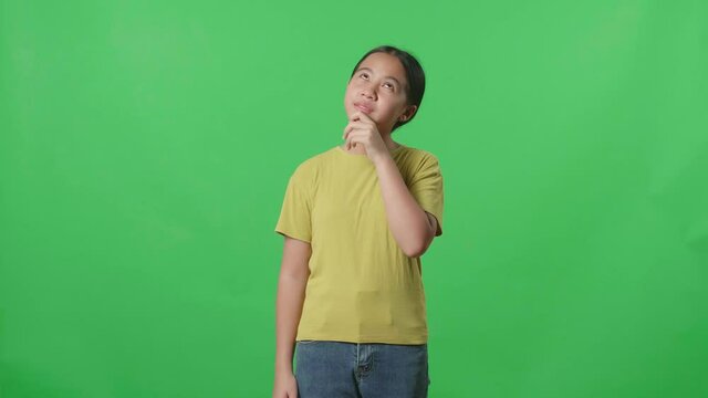 Young Asian Kid Girl Thinking About Something And Looking Around Then Raising Her Index Finger And Eyes Wide Open While Standing On Green Screen In The Studio

