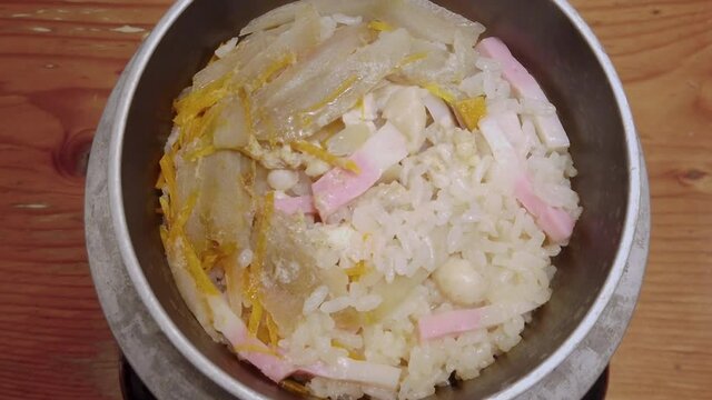 Close up video depicting a Japanese rice dish called Takikomi Gohan seasoned with dashi and soy sauce along with vegetables, kamaboko fish or bamboo shoots in a metal bowl.