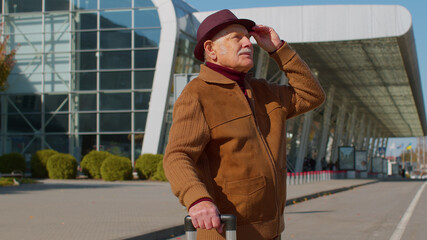 Fototapeta premium Portrait of elderly retired man tourist near airport terminal waiting boarding on plane for traveling. Stylish senior mature grandfather carrying luggage suitcases bag to railway station. Vacation