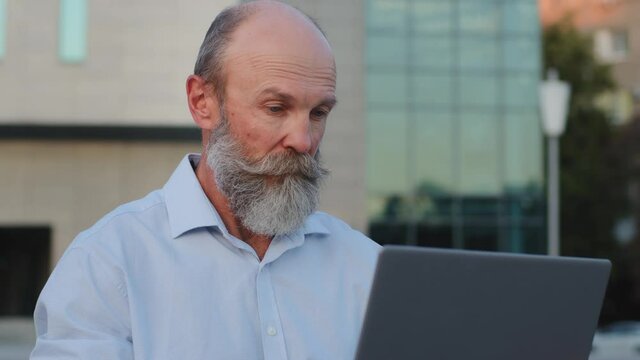Close-up pensive elderly man with gray beard sitting outdoors using laptop. Serious pensioner works remotely online. Old person mature specialist retirement age uses modern technology, computer app