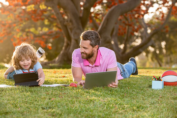 Happy child with father learning outdoor by studying online and working on laptop in green field.